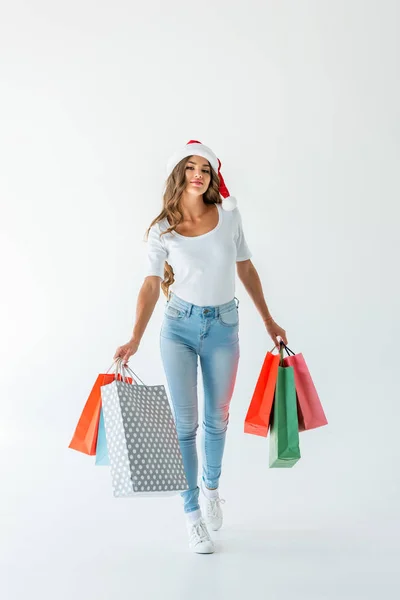 Приваблива щаслива дівчина в капелюсі Санта, що йде з сумками, ізольована на білому — стокове фото