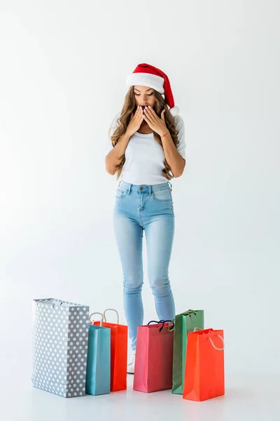 Chica sorprendida en sombrero de santa mirando bolsas de compras, aislado en blanco - foto de stock