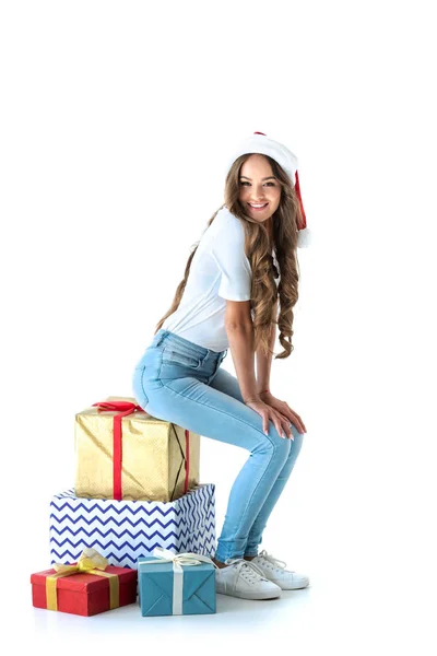 Belle fille souriante dans le chapeau de Père Noël assis sur les cadeaux de Noël, isolé sur blanc — Photo de stock