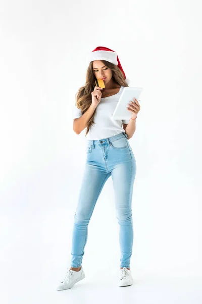 Pensive woman in santa hat shopping online with credit card and tablet, isolated on white — Stock Photo