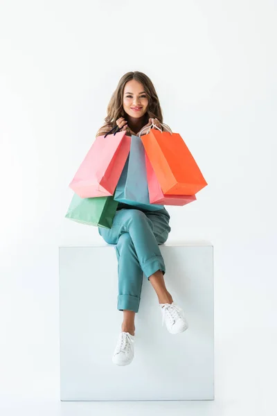 Sorridente shopaholic seduto su cubo bianco con borse della spesa, isolato su bianco — Foto stock