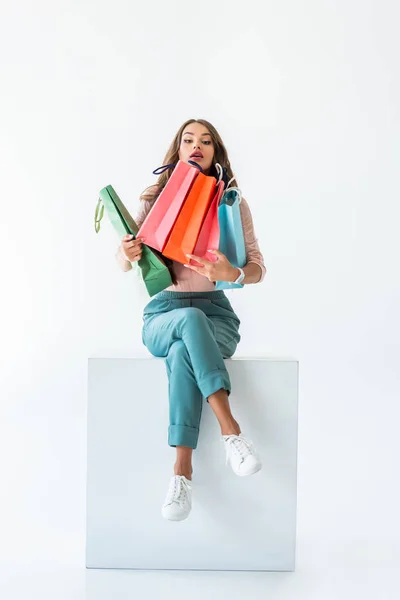 Schöne junge Shopperin sitzt auf White Cube mit Einkaufstaschen, isoliert auf Weiß — Stockfoto
