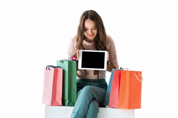 Улыбающаяся девушка с пакетами для покупок показывает планшет с пустым экраном, изолированный на белом — стоковое фото