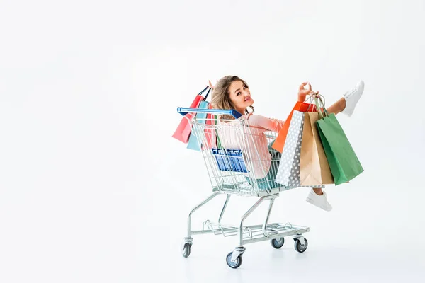 Beautiful happy girl with shopping bags sitting in shopping cart, isolated on white — Stock Photo
