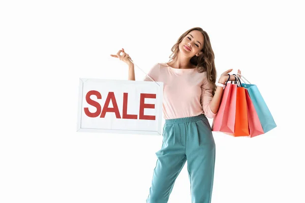 Bela menina sorridente segurando bordo com sinal de venda e sacos de compras, isolado no branco — Fotografia de Stock