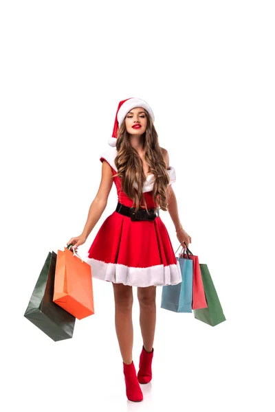 Atractiva chica morena en traje de santa claus sosteniendo bolsas de compras, aislado en blanco - foto de stock