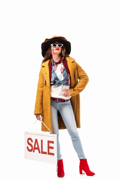 Jovem elegante em óculos de sol e roupa de outono com etiquetas de venda segurando placa com símbolo de venda, isolado em branco — Fotografia de Stock