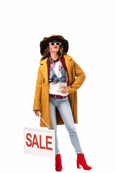 Mujer joven con estilo en gafas de sol y traje de otoño con etiquetas de la venta que sostiene el tablero con el símbolo de la venta, aislado en blanco - foto de stock