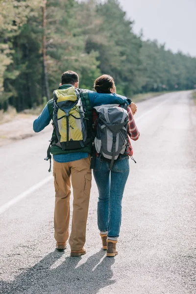 Visão traseira de casal de viajantes com mochilas andando na estrada — Fotografia de Stock