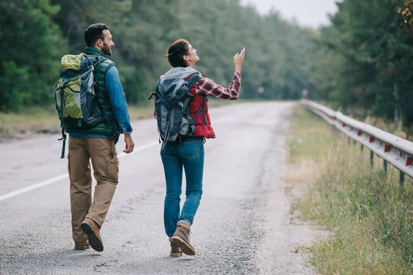 Vue arrière de couple de voyageurs avec sacs à dos marchant sur la route — Photo de stock