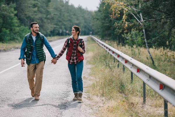 Couple of travelers with backpacks holding hands while walking on road — Stock Photo