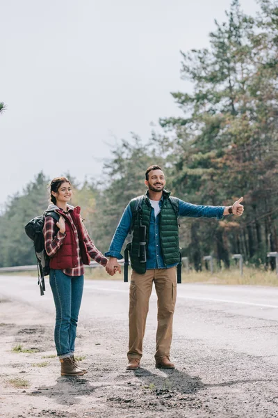 Pareja de viajeros con mochilas cogidas de la mano autostop en la carretera - foto de stock