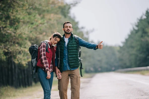 Couple of travelers with backpacks hitchhiking on road — Stock Photo