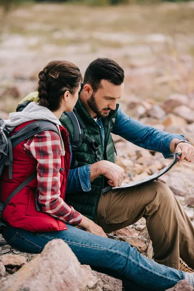 Seitenansicht von Wanderern, die auf der Karte das Ziel suchen, während sie am Sandstrand auf Felsen sitzen — Stockfoto