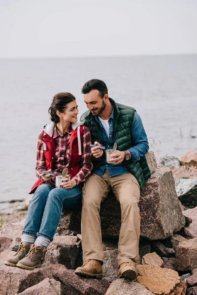 Lächelndes Paar mit Essen in Konservendosen auf Felsen am Sandstrand — Stockfoto