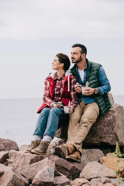 Nachdenkliches Paar von Reisenden mit Essen in Konservendosen, die sich auf Felsen am Sandstrand ausruhen — Stockfoto