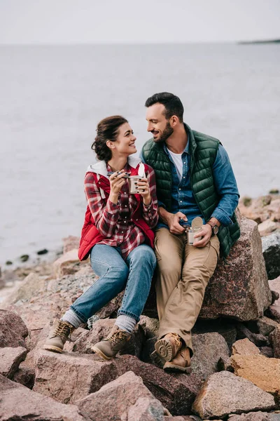Smiling couple of travelers eating food from cans while resting on rocks on sandy beach — Stock Photo