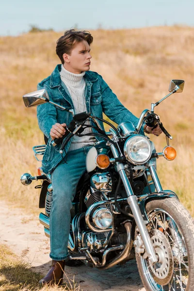 Schöner, stylischer junger Mann sitzt auf einem Oldtimer-Motorrad auf einer ländlichen Wiese — Stockfoto