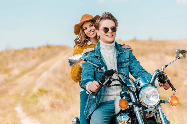 Jeune couple souriant assis sur moto rétro sur prairie rurale — Photo de stock