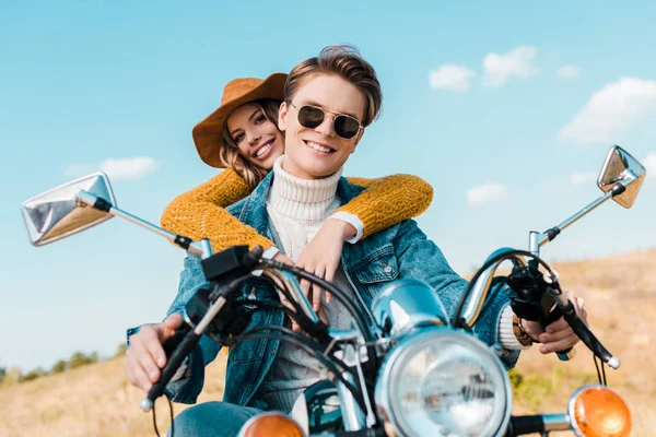 Novia atractiva abrazando novio sentado en moto retro en el prado - foto de stock