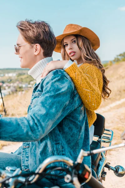 Beautiful girlfriend looking at camera while boyfriend sitting on motorbike on meadow — Stock Photo