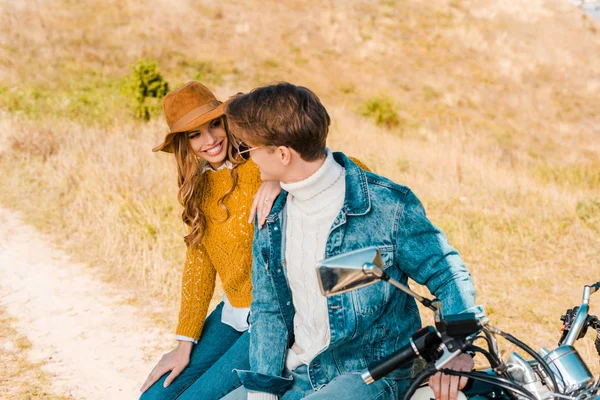 Улыбающаяся пара, сидящая на мотоцикле и смотрящая друг на друга на лугу — стоковое фото