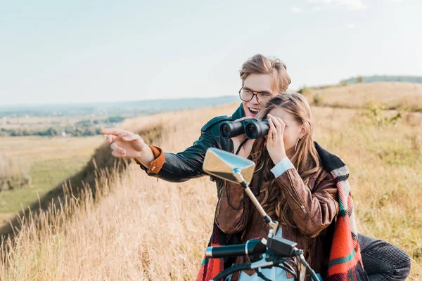 Girlfriend looking through binoculars near smiling boyfriend showing on something — Stock Photo