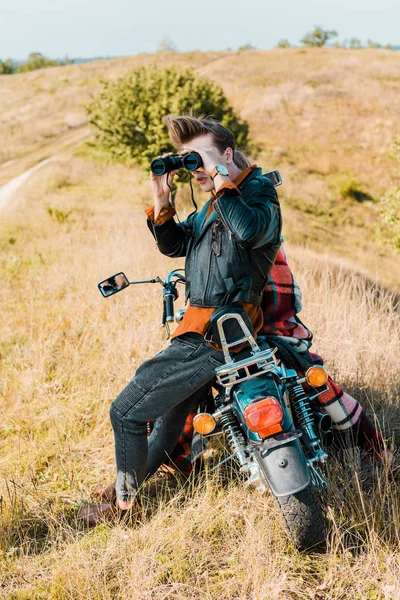 Junger gutaussehender Mann mit Fernglas sitzt auf Oldtimer-Motorrad auf bäuerlicher Wiese — Stockfoto