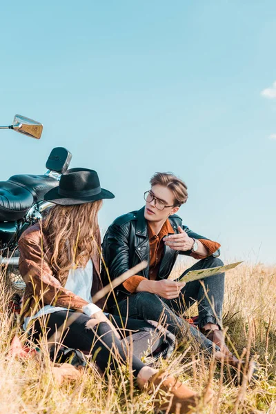 Young boyfriend in glasses holding map and looking at beautiful girlfriend sitting on meadow — Stock Photo