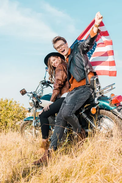 Couple heureux avec drapeau américain assis sur prairie, concept de fête de l'indépendance — Photo de stock