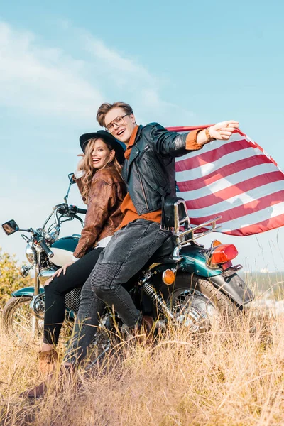 Молодий посміхаючись пара з американським прапором, сидячи на мотоциклі, День незалежності концепції — стокове фото