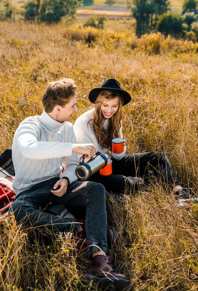 Happy couple pouring hot drink from thermos and resting on rural meadow — Stock Photo