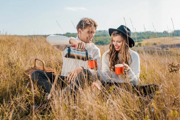 Happy boyfriend pouring hot drink from thermos into cups for girlfriend on rural meadow — Stock Photo