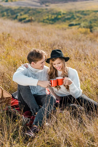 Joven sonriente pareja tintineo con tazas y relajarse en el prado rural - foto de stock