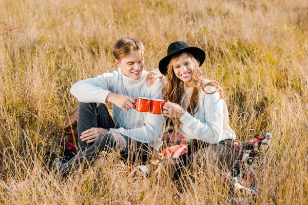Молодая пара, звенящая с чашками и сидящая на клетке на сельском лугу — стоковое фото