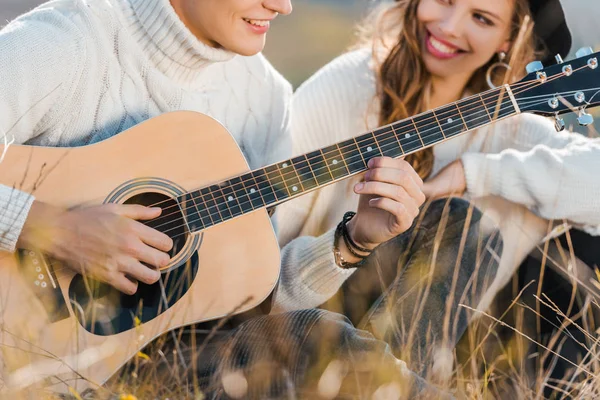 Обрезанный вид улыбающейся пары, расслабляющейся на лугу, пока молодой человек играет на гитаре — стоковое фото