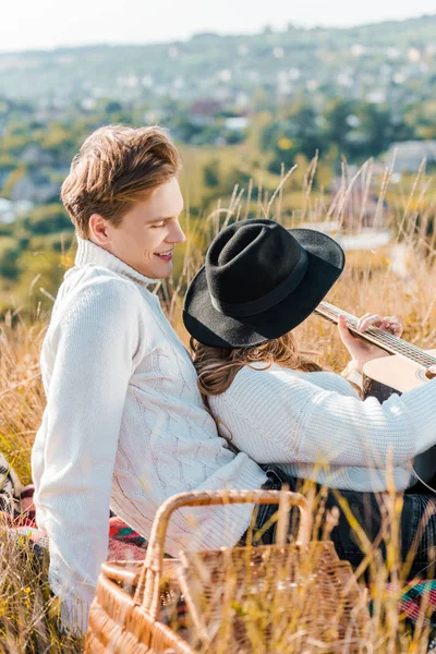 Namorada de chapéu preto segurando guitarra enquanto namorado relaxando na grama — Fotografia de Stock