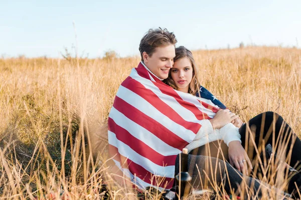 Sonriente pareja con bandera americana relajándose en prado rural, concepto del día de la independencia - foto de stock