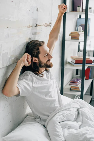Вид сбоку молодого бородатого мужчины с длинными волосами, делающего растяжку в утреннее время в постели дома — стоковое фото