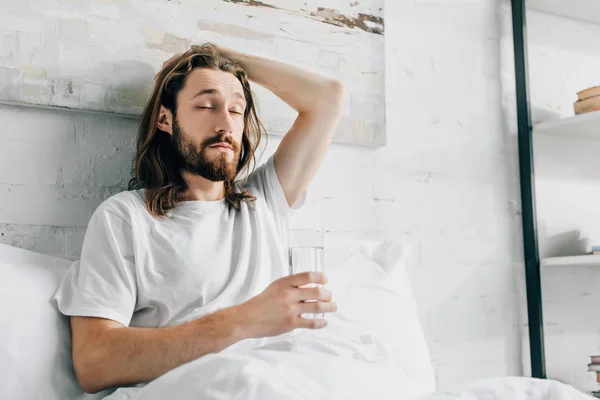 Jovem barbudo homem com cabelos longos tendo ressaca e tomando pílula com vidro de água no quarto em casa — Fotografia de Stock