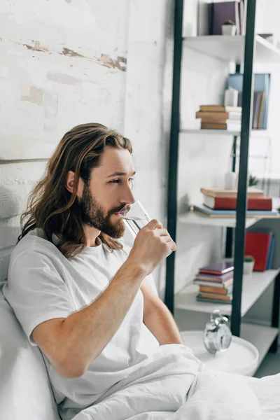 Side view of young bearded man with long hair having headache and taking pill with glass of water in bedroom at home — Stock Photo