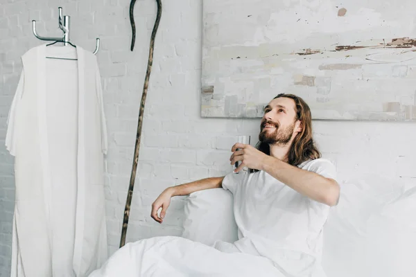 Happy Jesus looking up and holding glass of water in bed during morning time at home — Stock Photo