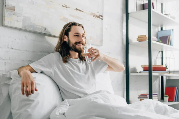 Улыбающийся бородатый мужчина с длинными волосами пьет воду в спальне дома — стоковое фото