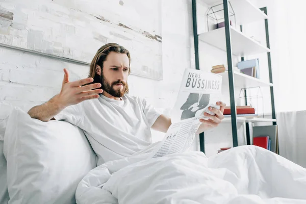 Сердитый бородатый бизнесмен с длинными волосами читает газету и жестикулирует вручную в постели в утреннее время дома — стоковое фото