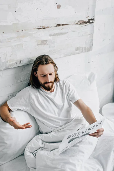 Vue grand angle de l'homme d'affaires barbu émotionnel avec les cheveux longs gesticulant à la main en lisant le journal dans le lit pendant le matin à la maison — Photo de stock