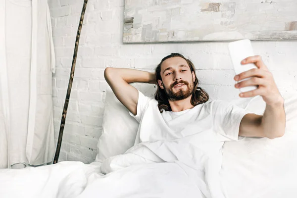 Улыбаясь Иисус делает селфи в постели в утреннее время дома — стоковое фото
