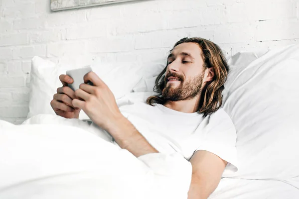 Jeune homme barbu aux cheveux longs en utilisant un smartphone au lit le matin à la maison — Photo de stock