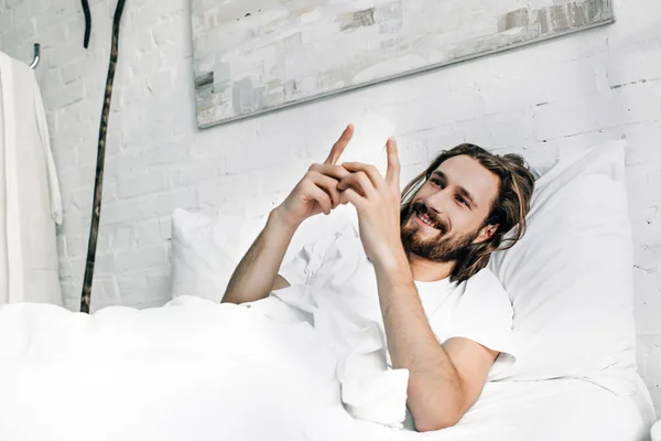 Sorrindo Jesus usando smartphone na cama durante o horário da manhã em casa — Fotografia de Stock