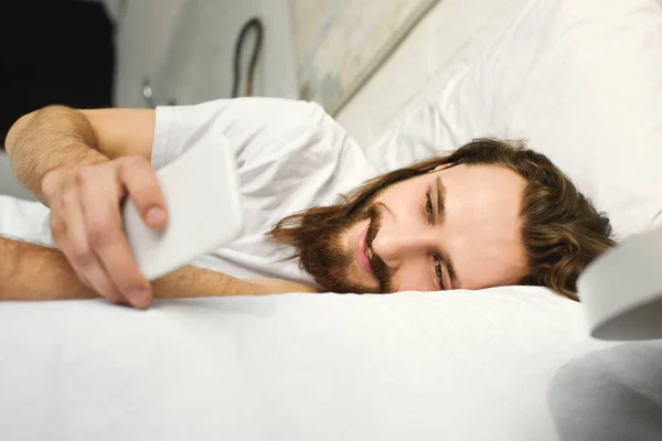 Улыбающийся бородатый мужчина с длинными волосами с помощью смартфона в постели в утреннее время дома — стоковое фото
