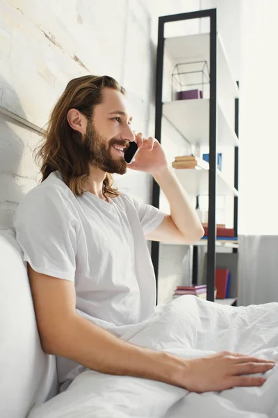 Вид сбоку улыбающегося бородатого мужчины с длинными волосами, говорящего по смартфону в постели в утреннее время дома — стоковое фото